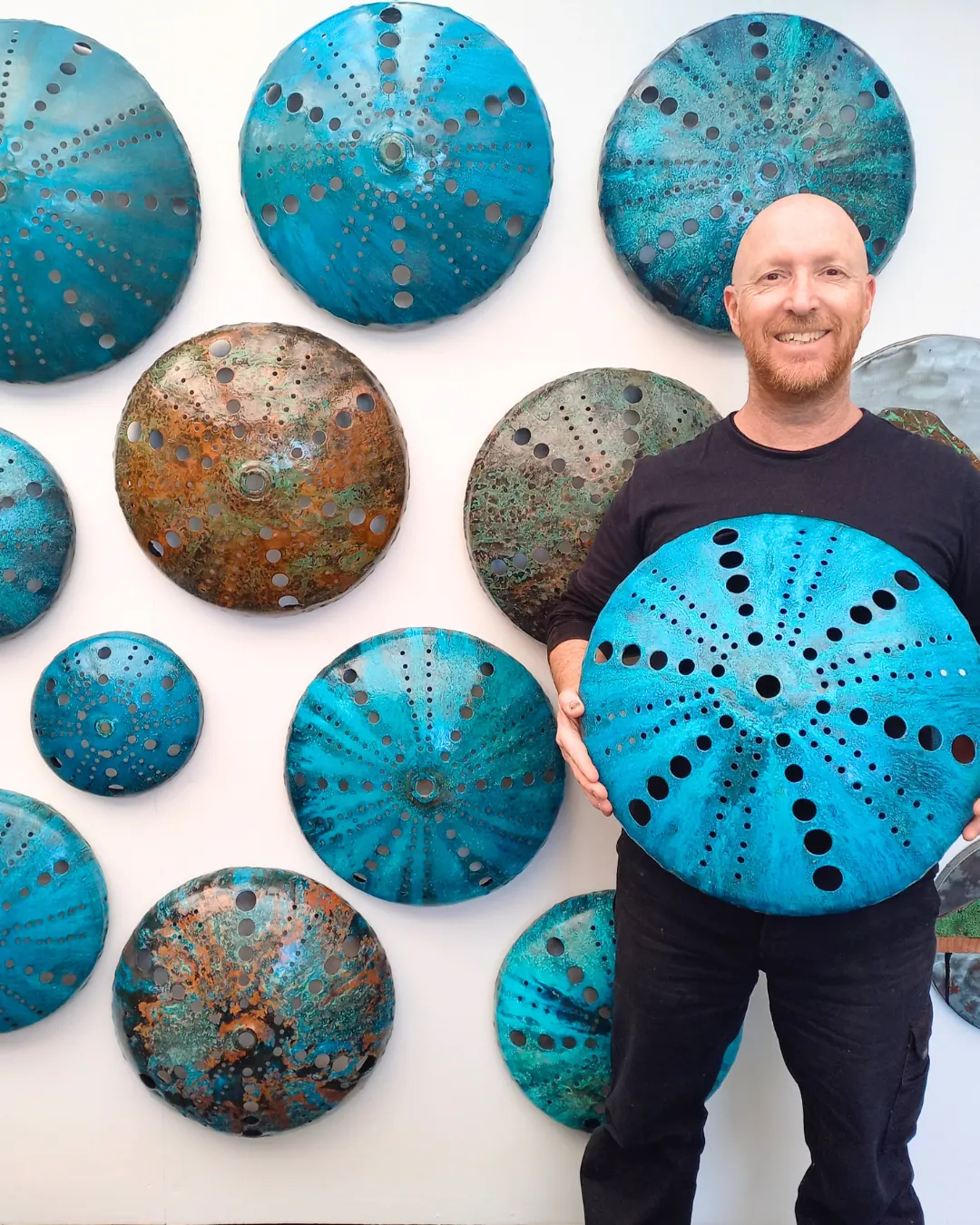 Isaac Weston holding a blue-patinated copper kina in front of a wall of his sea urchin works